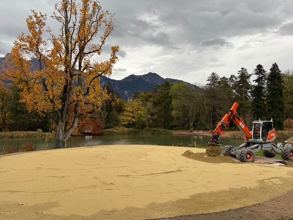 Ein Bagger schüttet Sand an einem Seeufer unter bewölktem Himmel auf, umgeben von herbstlichen Bäumen und fernen Bergen, und vermittelt so ein Bild von Ruhe und Fleiß.
