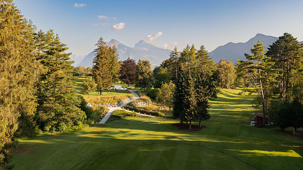 Ein malerischer Golfplatz, umgeben von hohen Bäumen und Bergen unter einem klaren blauen Himmel. Das Sonnenlicht wirft sanfte Schatten und schafft eine ruhige und einladende Atmosphäre.
