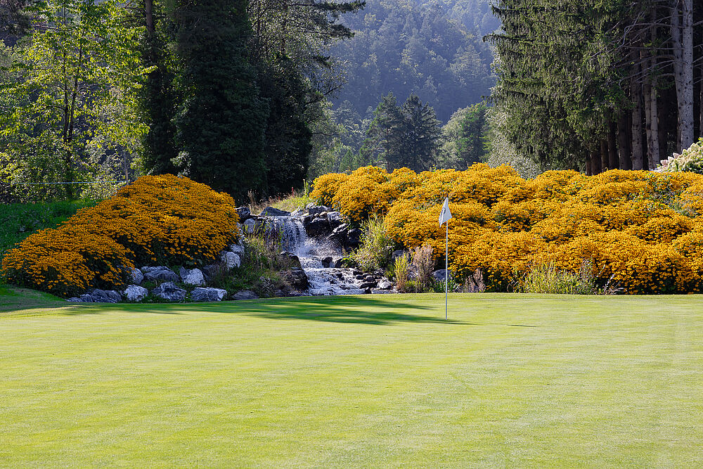 Eine Szene auf Green 11 mit leuchtend orangefarbenen Blumen, grünen Sträuchern und einem kleinen Wasserfall, der sich über Felsen ergiesst, vor einem Waldhintergrund und einem klaren blauen Himmel.