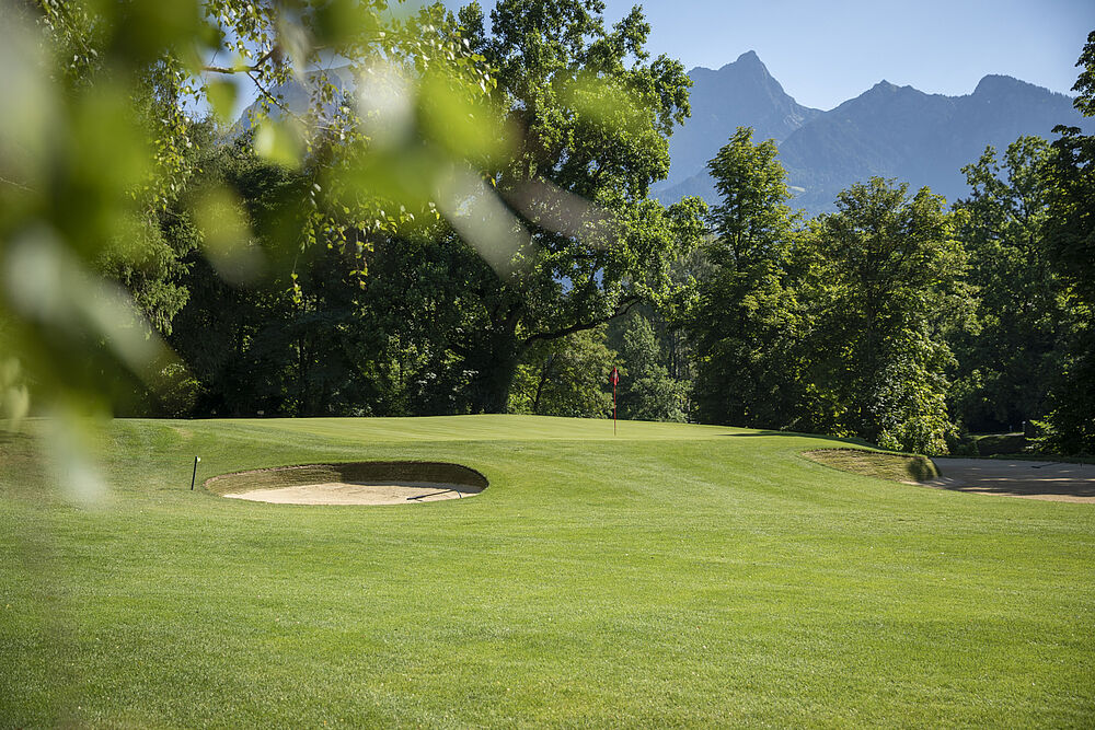 Ein grüner Golfplatz mit Sandbunkern, umgeben von Bäumen und vor der Kulisse der fernen Berge unter einem klaren blauen Himmel.