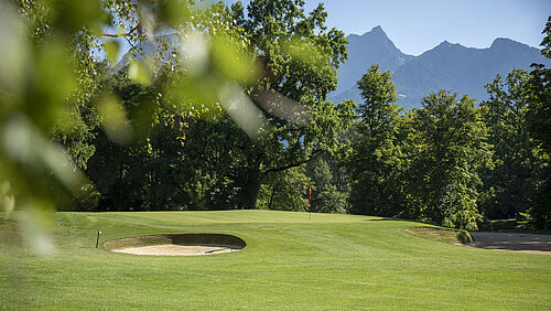 A green golf course with sand bunkers, surrounded by trees and set against the backdrop of distant mountains under a clear blue sky.