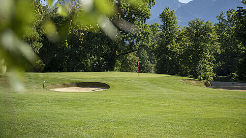 Ein grüner Golfplatz mit Sandbunkern, umgeben von Bäumen und vor der Kulisse der fernen Berge unter einem klaren blauen Himmel.