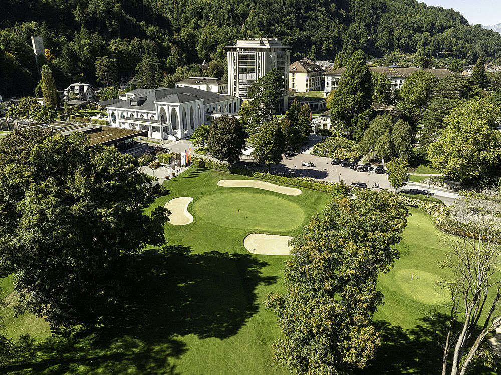 Luftaufnahme des Golfplatzes, umgeben von üppigen Bäumen und dem Grand Resort Bad Ragaz. Die Szene ist ruhig, das Sonnenlicht wirft sanfte Schatten.