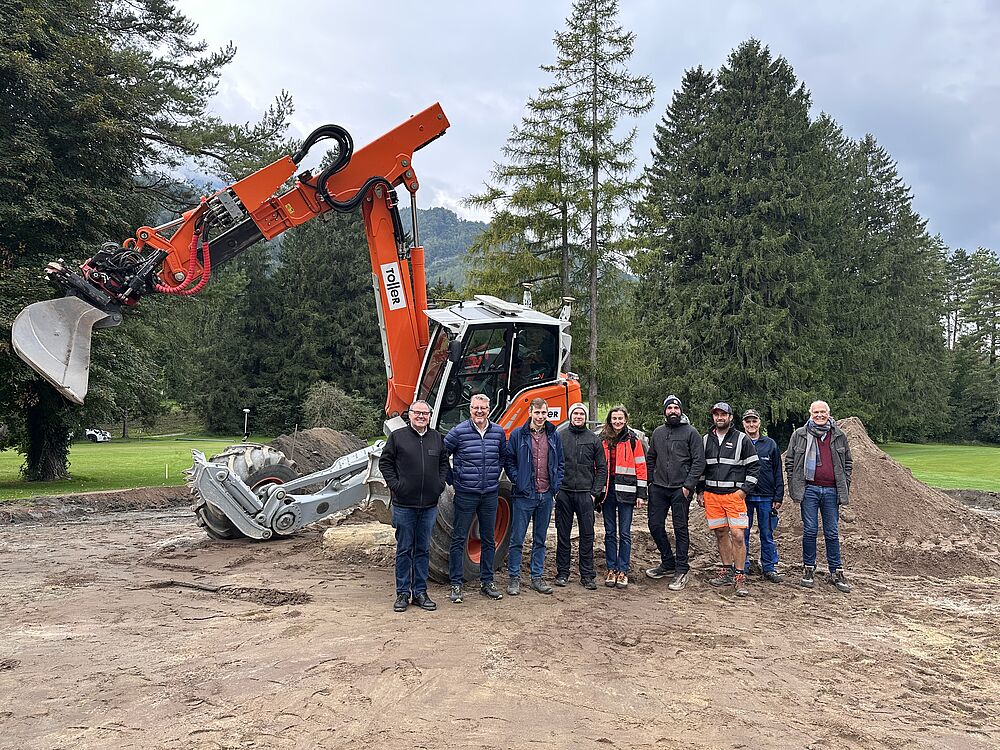 Eine Gruppe von neun Personen steht auf einer Baustelle vor einem orangefarbenen Bagger mit einer großen Schaufel. Im Hintergrund sind hohe Bäume und Berge zu sehen, über ihnen ein bewölkter Himmel. Die Stimmung ist kooperativ.