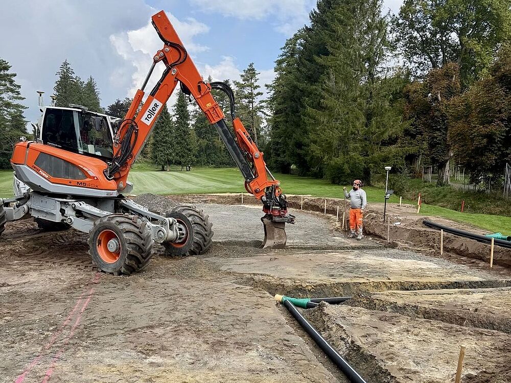 Eine Baustelle mit einem orangefarbenen Bagger auf Kies, der Land rodet. Arbeiter und verstreute Rohre sind zu sehen, umgeben von Bäumen unter einem teilweise bewölkten Himmel.