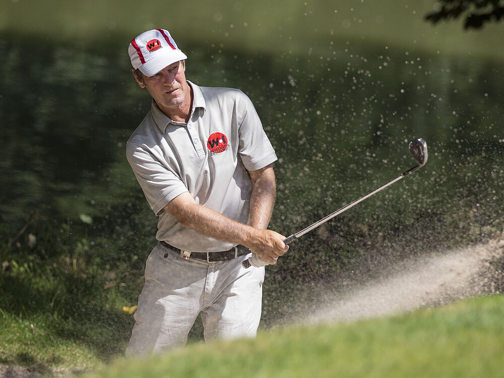 Ein Golfer in grauem Outfit und roter Mütze schlägt aus einem Sandbunker ab, wobei Sand umherfliegt. Die Szene vermittelt Konzentration und Präzision.