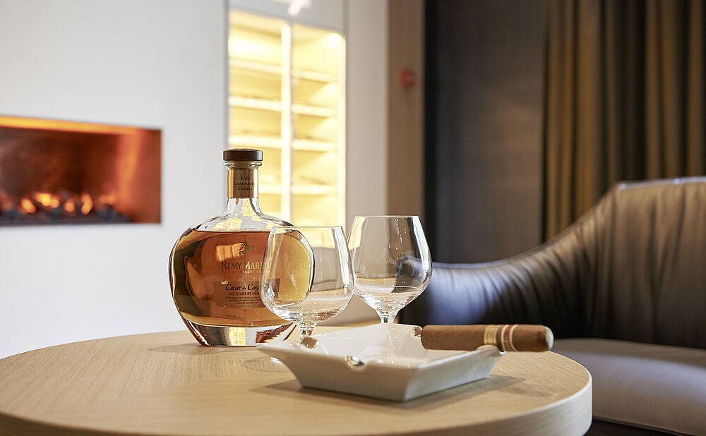 A cozy room with a glowing fireplace. A round table holds a bottle of liquor, two empty glasses, and a cigar on a tray, creating a warm, inviting atmosphere.