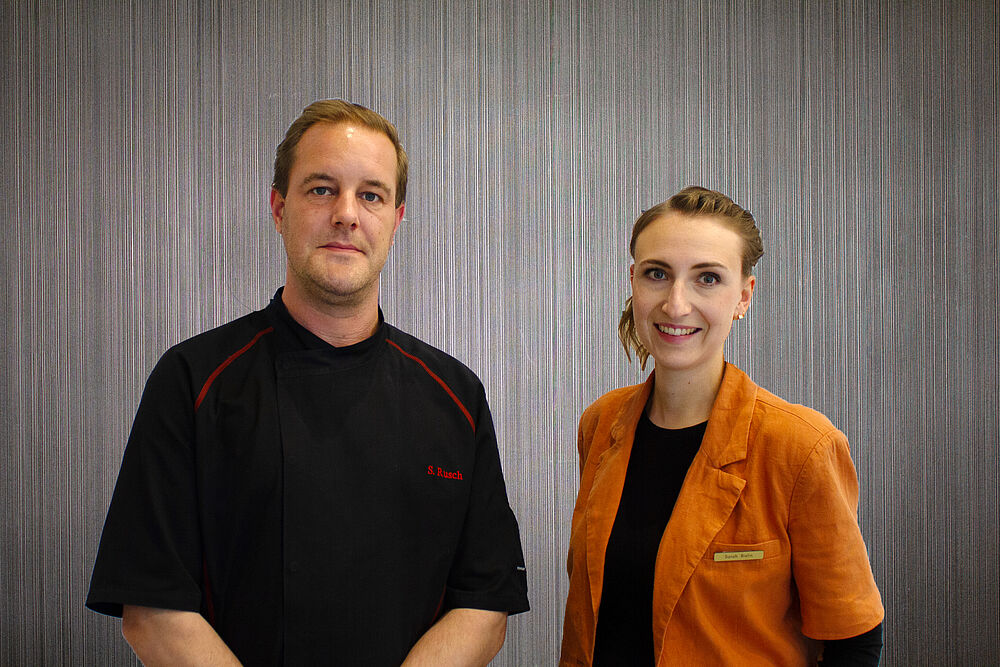 Simon Rusch in einem schwarzen Kochshirt und Sarah Bislin mit einem schwarzen T-Shirt und orangenem Blazer.