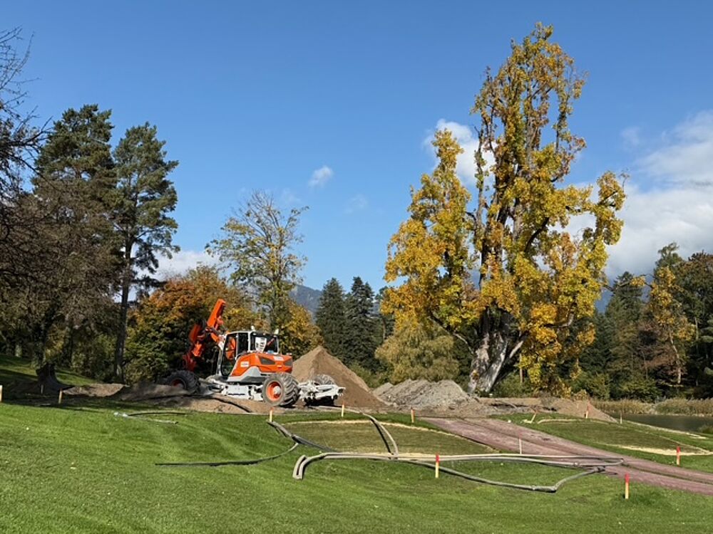Eine Baustelle in einem Park mit einem weiß-orangefarbenen Bagger zwischen Erdhaufen. Hohe Herbstbäume und ein strahlend blauer Himmel bilden eine ruhige Kulisse.
