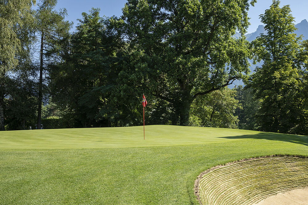 Ein gepflegter Golfplatz mit üppigem grünem Gras, einem Sandbunker auf der rechten Seite und einer roten Flagge, die das Loch markiert. Im Hintergrund hohe Bäume und Berge.