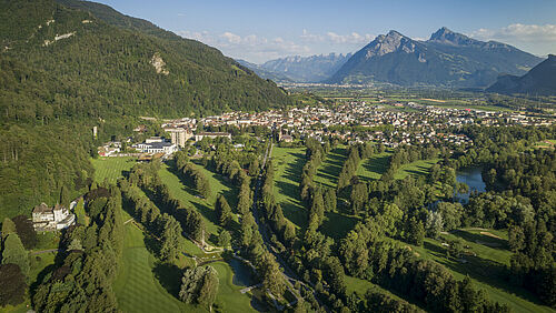 Luftaufnahme eines Golfplatzes mit sattgrünen Fairways, umgeben von dichten Wäldern und Bad Ragaz, vor dem Hintergrund der fernen Berge.