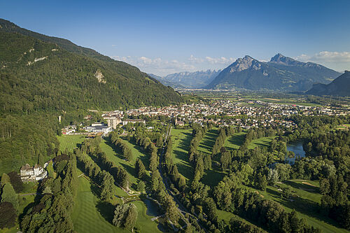 Aerial view of a golf course with lush green fairways, surrounded by dense forests and Bad Ragaz, with the distant mountains as a backdrop.