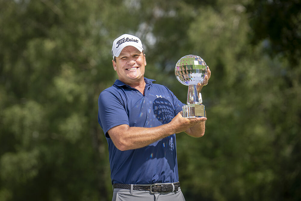 Der Sieger vom Swiss Seniors Open 2024, Jarmo Sandelin, hält den Glaspokal in die Luft. Er lächelt, trägt eine weisse Mütze und ein blaues Golfshirt.