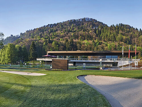 Das moderne Clubhaus Haus mit großen Fenstern und Terrasse, eingebettet in eine grüne Landschaft direkt hinter Abschlag 1. Die Berge und der blaue Himmel bilden eine heitere Kulisse, die Ruhe ausstrahlt.