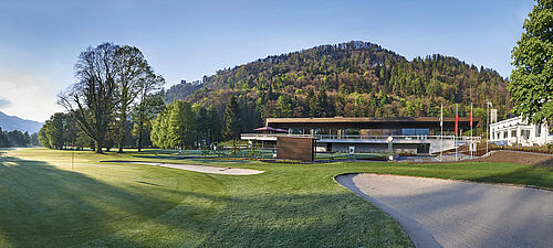 The modern clubhouse with large windows and terrace is nestled in a green landscape directly behind tee 1. The mountains and the blue sky form a serene backdrop that radiates tranquillity.