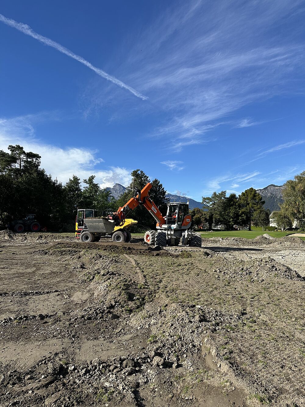 Ein leuchtend orangefarbener Bagger arbeitet unter strahlend blauem Himmel auf einer Baustelle. Bäume und ferne Berge rahmen die Szene ein und vermitteln Produktivität.