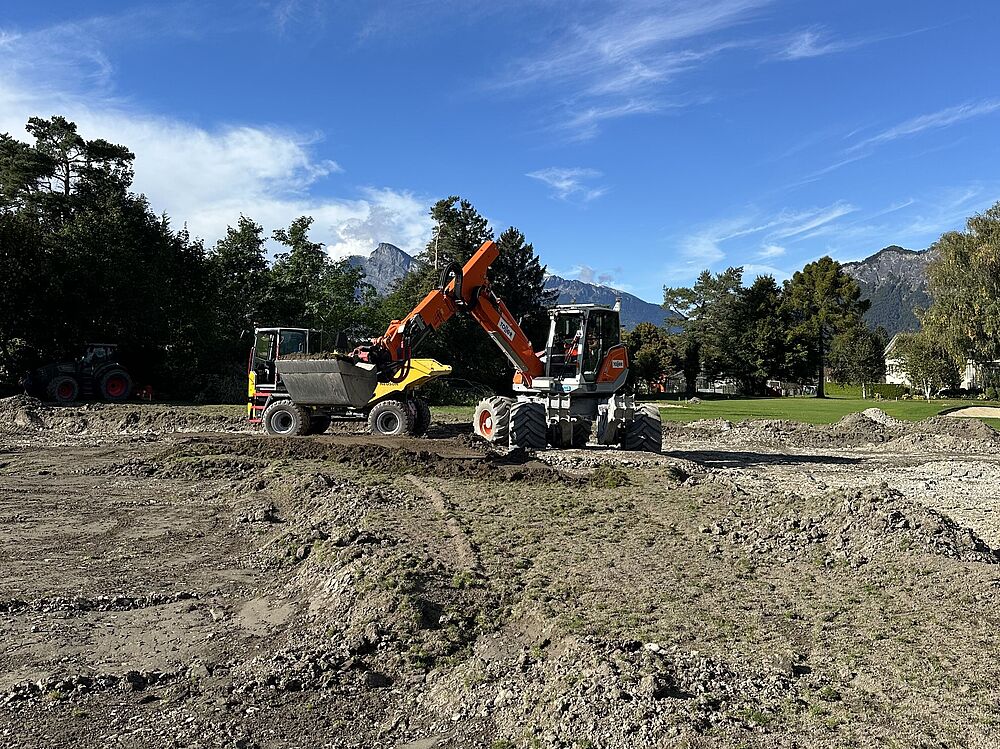 Ein leuchtend orangefarbener Bagger arbeitet unter strahlend blauem Himmel auf einer Baustelle. Bäume und ferne Berge rahmen die Szene ein und vermitteln Produktivität.
