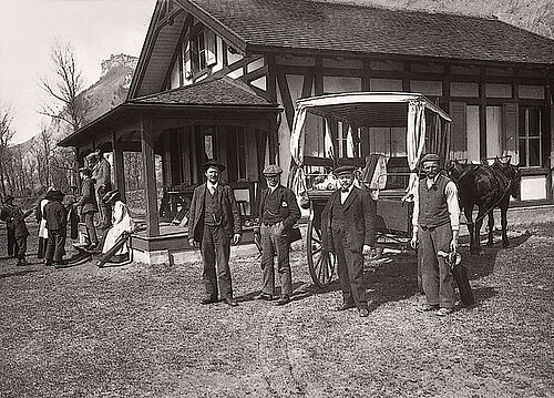 Ein Bild in schwarz und weiss. Sie sitzen vor einem Gebäude, das alte Clubhaus und unterhalten sich. Rechts im Bild steht eine Pferdekutsche und daneben zwei Männer chiq gekleidet und mit Hut.
