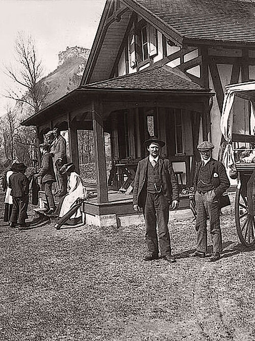 A picture in black and white. They are sitting in front of a building, the old clubhouse, chatting. On the right of the picture is a horse-drawn carriage and next to it two men dressed in chiq and wearing hats.