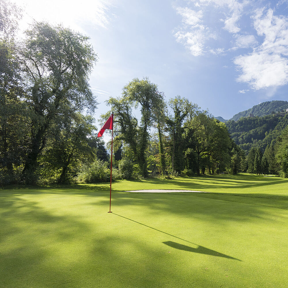 Ein sonnenbeschienener Golfplatz mit einer roten Flagge auf dem Grün, umgeben von üppigen Bäumen und Bergen unter einem strahlend blauen Himmel mit vereinzelten Wolken.