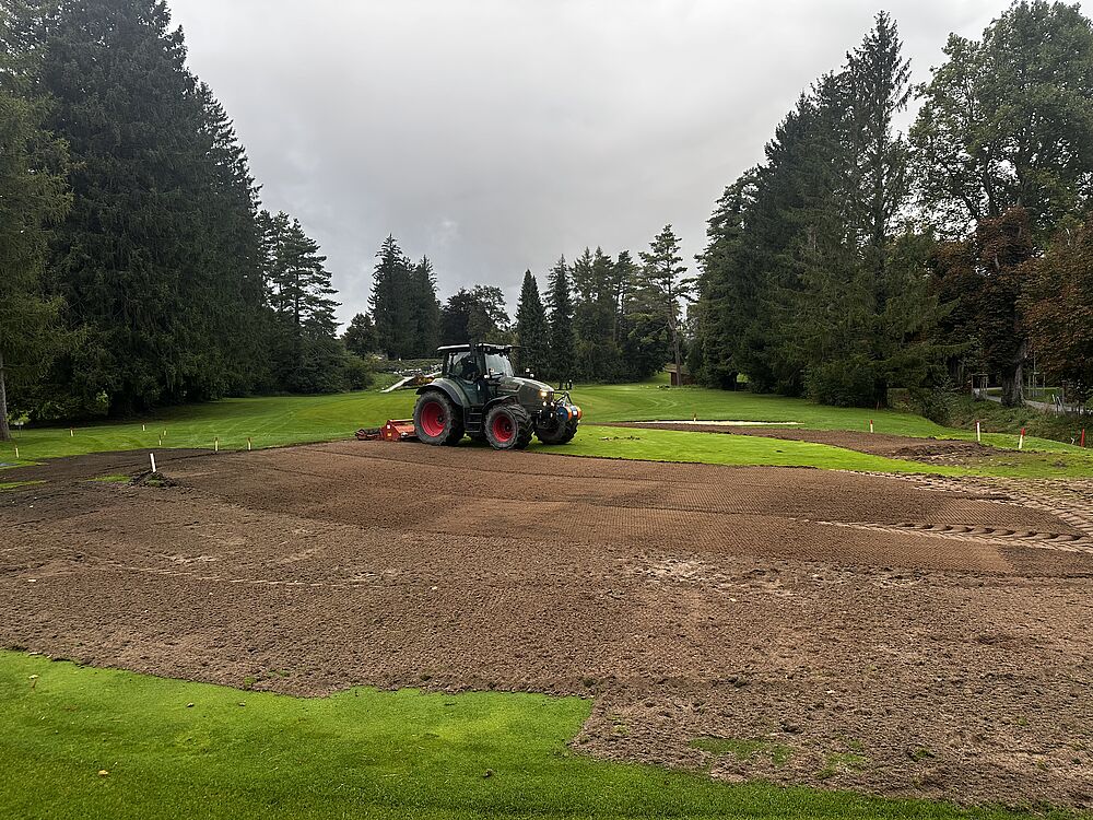 Ein Traktor mit roten Rädern pflügt ein großes, schlammiges Feld in einem üppig grünen Park, umgeben von hohen Bäumen unter einem bewölkten Himmel.