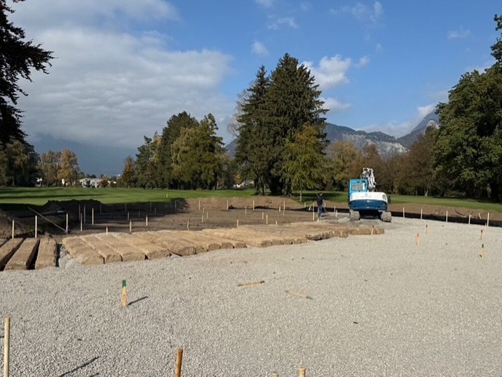 Eine Baustelle mit Kies und Holzplanken liegt vor einer malerischen Kulisse aus Bäumen und Bergen. Ein blauer Bagger steht unter einem teilweise bewölkten Himmel auf dem Gelände.
