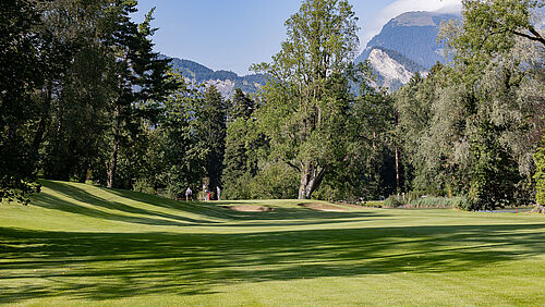 Der Golfplatz bei Loch 14 mit hohen Bäumen unter blauem Himmel. Das Sonnenlicht fällt durch die Blätter und schafft eine ruhige und friedliche Atmosphäre. Im Vordergrund zwei Bunker und rechts vom Bild der Giessensee.