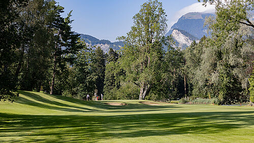 The golf course at hole 14 with tall trees under a blue sky. The sunlight falls through the leaves and creates a calm and peaceful atmosphere. Two bunkers in the foreground and Lake Giessen to the right of the picture.
