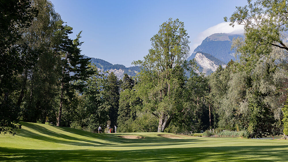 Der Golfplatz bei Loch 14 mit hohen Bäumen unter blauem Himmel. Das Sonnenlicht fällt durch die Blätter und schafft eine ruhige und friedliche Atmosphäre. Im Vordergrund zwei Bunker und rechts vom Bild der Giessensee.