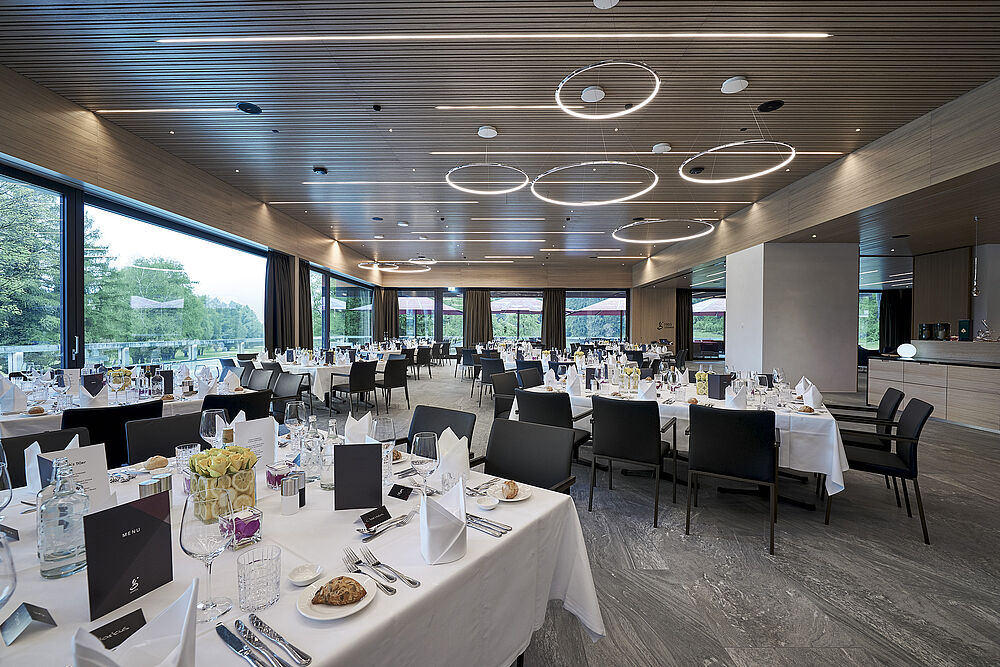 Modern restaurant with large windows, elegantly laid tables with white tablecloths and black chairs. Soft ceiling lighting creates a sophisticated ambience.
