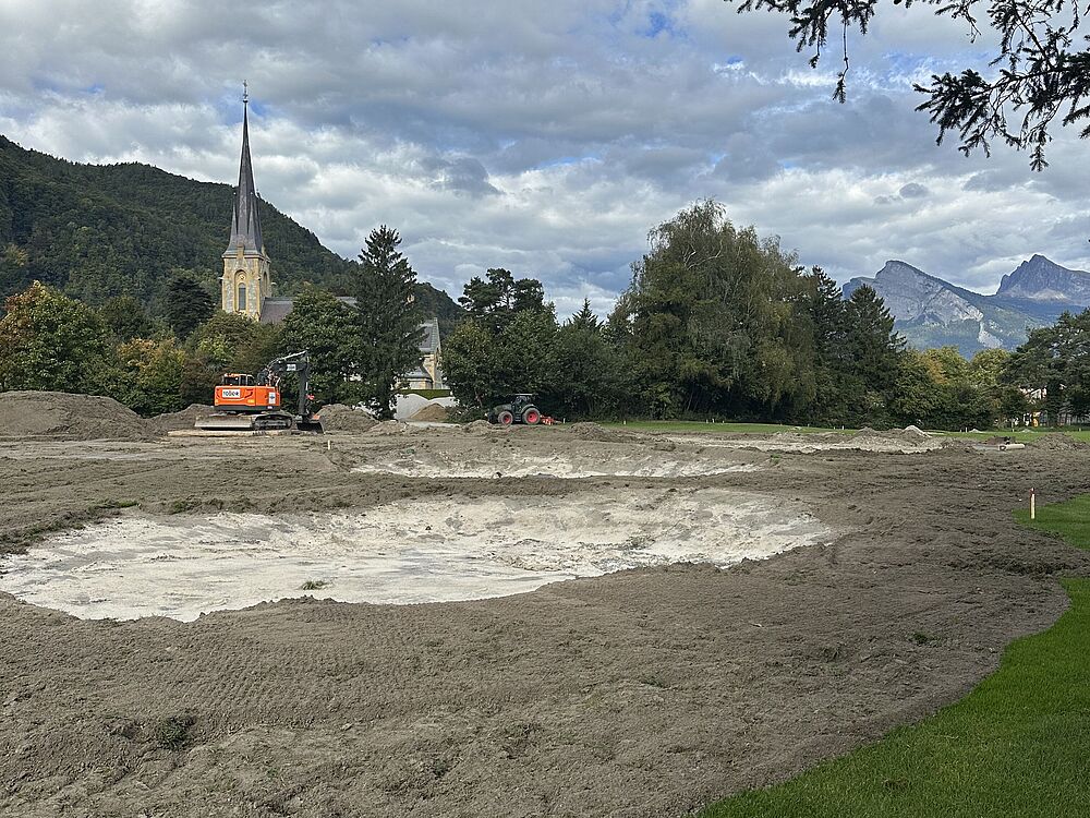 Eine Wiese mit einer großen Ausgrabungsstätte im Vordergrund. Vor dem Hintergrund von Bergen und einem bewölkten Himmel ist eine Kirche mit einem hohen Turm zu sehen.