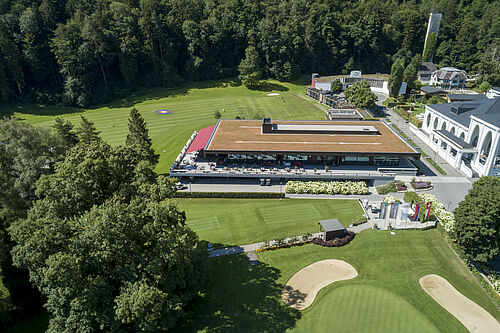 Luftaufnahme eines modernen Golfclubhauses mit grünem Dach, umgeben von üppigen Bäumen und einem gepflegten Golfplatz. Die Umgebung ist ruhig und einladend.