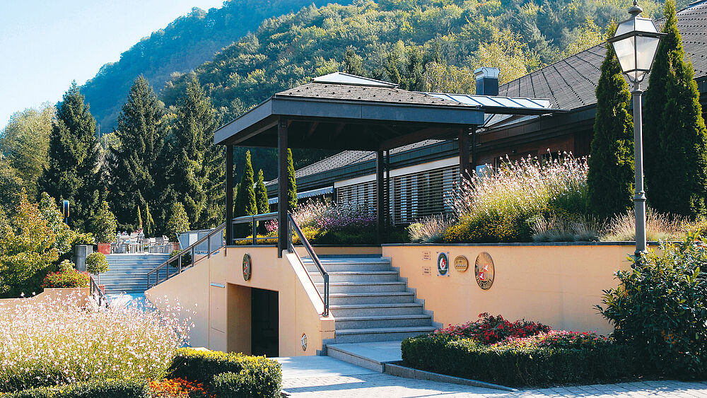 Das Bild zeigt das alte Clubhaus des Golf Club Bad Ragaz. Ein elegantes Gebäude mit Garten in Hanglage, Treppen, die nach oben führen, und üppigem Grün drum herum. Im Hintergrund die malerischen Berge unter einem klaren blauen Himmel. Friedliche Atmosphäre.