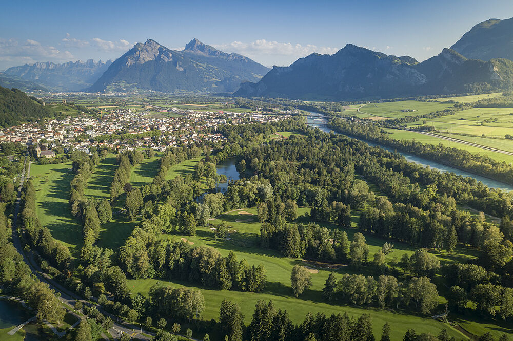 Luftaufnahme des Golfplatzes mit Wäldern, einem sich schlängelnden Fluss und einer kleinen Stadt, die sich unter einem klaren blauen Himmel an die Berge schmiegt.