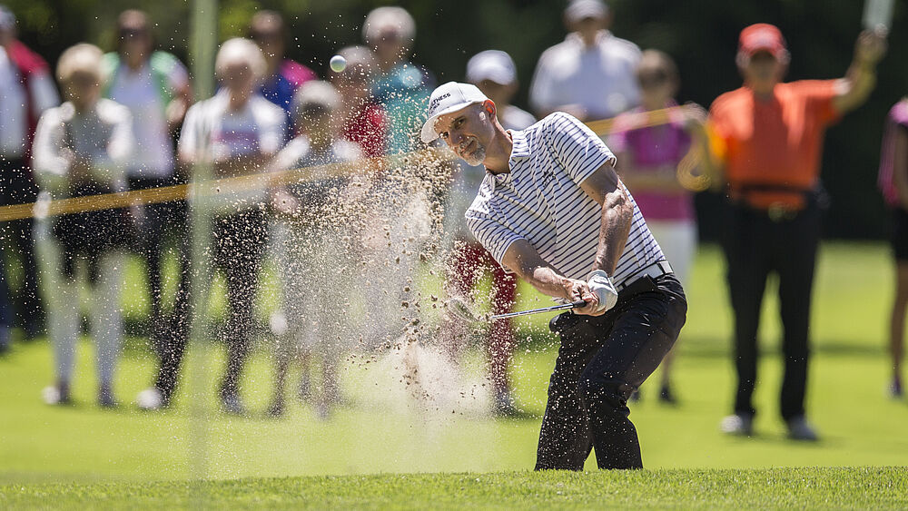 Golfer, der an einem sonnigen Tag einen Sandschlag macht, wobei der Sand aus dem Bunker fliegt. Die Zuschauer im Hintergrund schauen aufmerksam zu. Fokussierte und dynamische Szene.