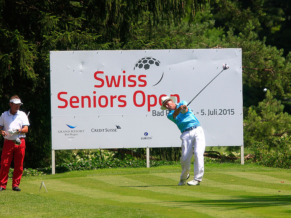Golfer im Schwung bei den Swiss Seniors Open, mit einem großen Veranstaltungsschild und üppigem Grün im Hintergrund, das Konzentration und Präzision vermittelt.