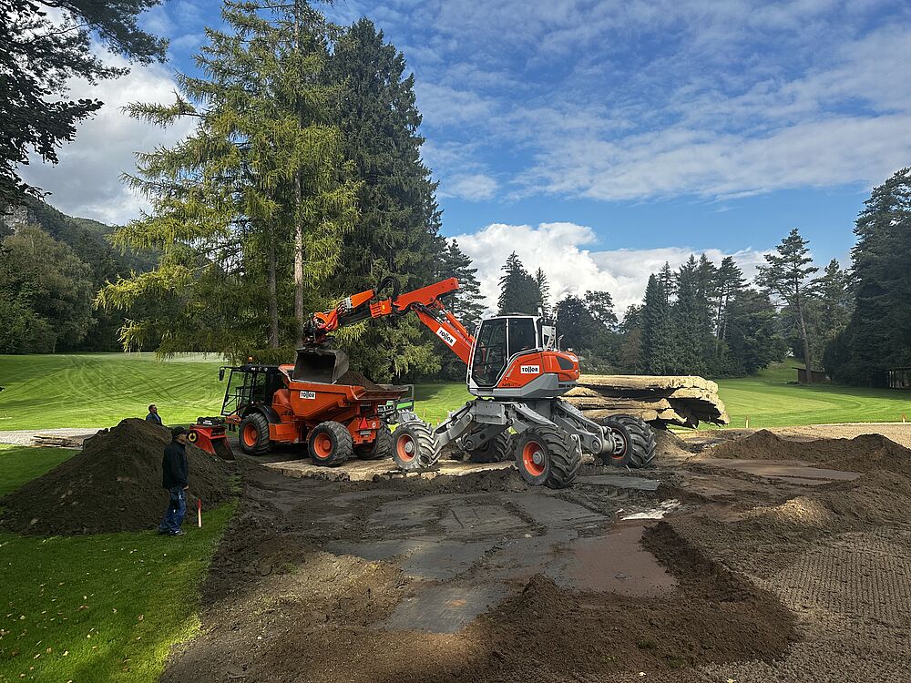 Baustelle auf einer Wiese mit zwei leuchtend orangefarbenen Baggern, die Erde bewegen. Hohe Bäume und ein bewölkter Himmel bilden den Hintergrund. Arbeiter stehen in der Nähe.