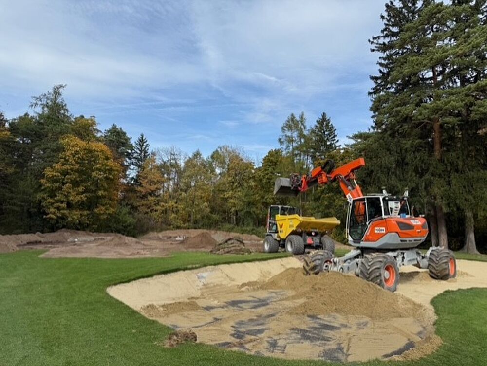 Ein Baufahrzeug lädt Sand in einen Golfplatzbunker, der von üppigem Gras und herbstlichen Bäumen umgeben ist, unter einem teilweise bewölkten Himmel, was den Fortschritt verdeutlicht.
