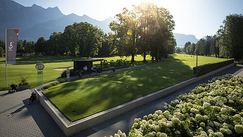 Sonnenbeschienener Park mit üppigem grünen Gras und verstreuten Bäumen, Berge im Hintergrund. Ein kleiner Pavillon und eine Fahne sind in der Szene zu sehen, die Ruhe ausstrahlen.