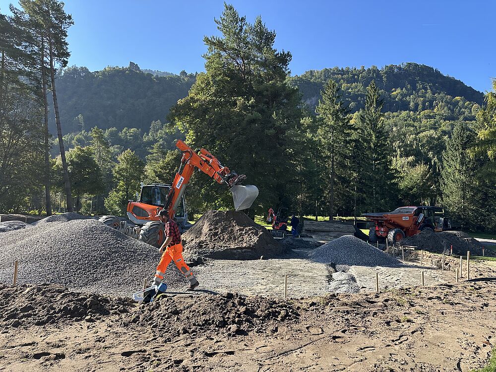 Eine Baustelle in einem Waldgebiet mit einem Arbeiter in leuchtend orangefarbener Kleidung, der neben einem kleinen Bagger entlanggeht. Im Hintergrund sind Kies- und Erdhaufen zu sehen, umgeben von hohen Bäumen und Bergen unter einem klaren blauen Himmel. Die Szene vermittelt ein Gefühl von Fleiß in einer natürlichen Umgebung.