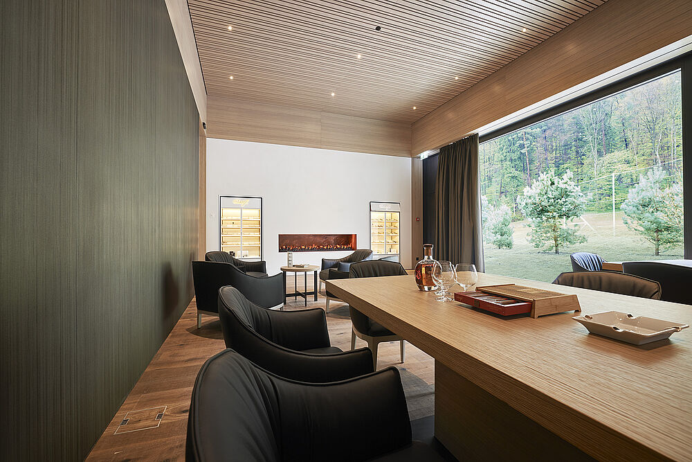 Die Smokers Loung im Clubhaus ist ein modernes Zimmer mit einem langen Holztisch, schwarzen Stühlen und einer dekorativen Decke. Ein grosses Fenster zeigt auf die Driving Range, die eine ruhige Atmosphäre schafft.