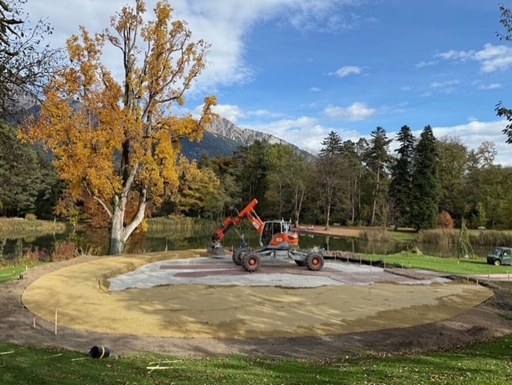 Ein Baufahrzeug arbeitet auf einer kreisförmigen Sandfläche, die von Gras und Bäumen mit Herbstlaub umgeben ist. Im Hintergrund sind Berge und ein See unter blauem Himmel zu sehen.