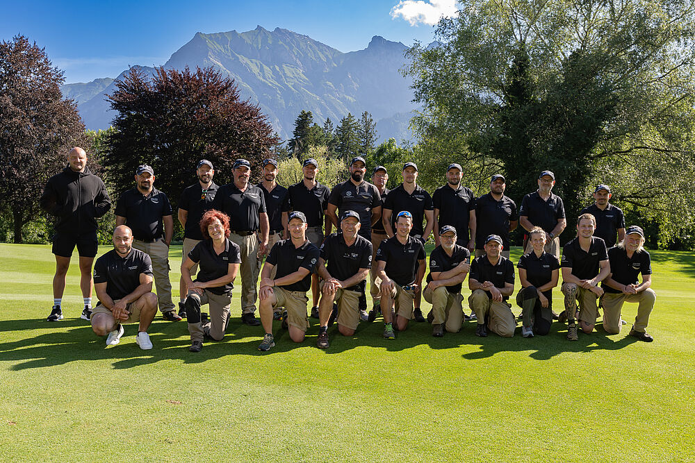 Das Greenkeeper Team bestehend aus 23 Personen. Alle tragen schwarze Tshirts und helle hosen. Sie stehen auf dem Fairway von Loch 14 und hinter ihnen sind die Berge zu sehen.