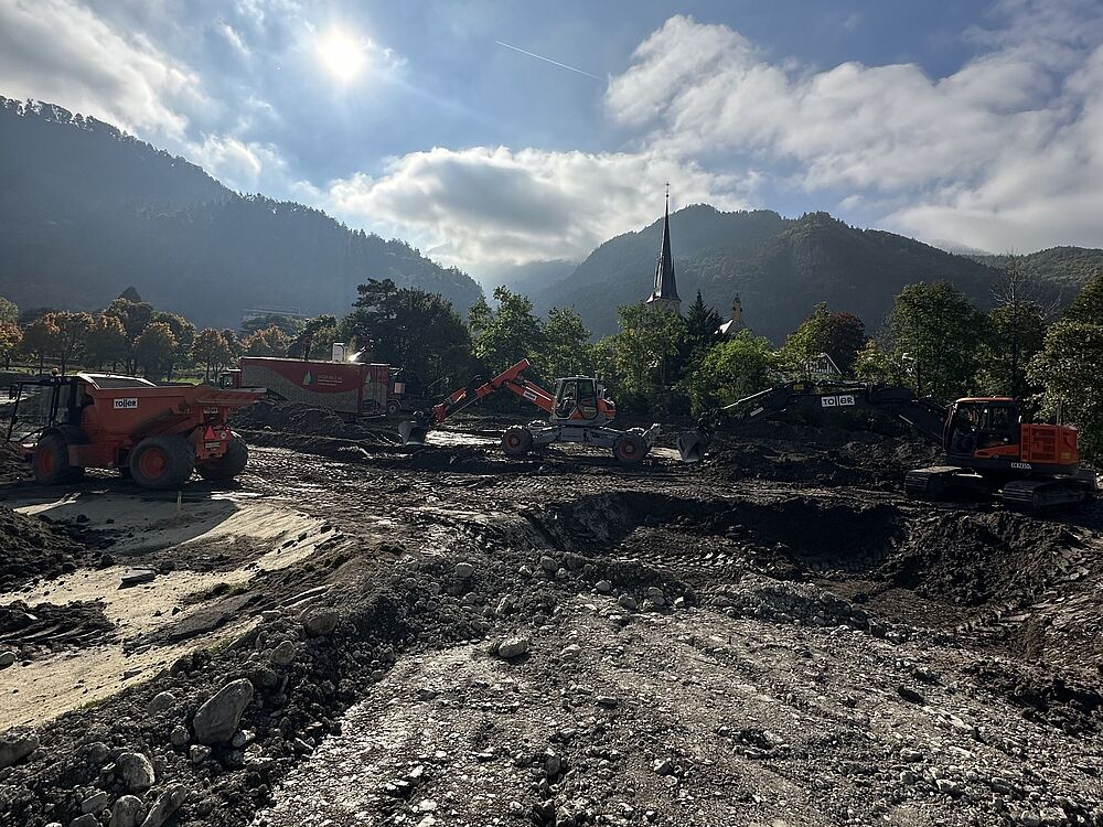 Baustelle mit Maschinen auf felsigem Boden unter strahlendem Himmel. Im Hintergrund Berge und Bäume, die eine dynamische und geschäftige Szene schaffen.