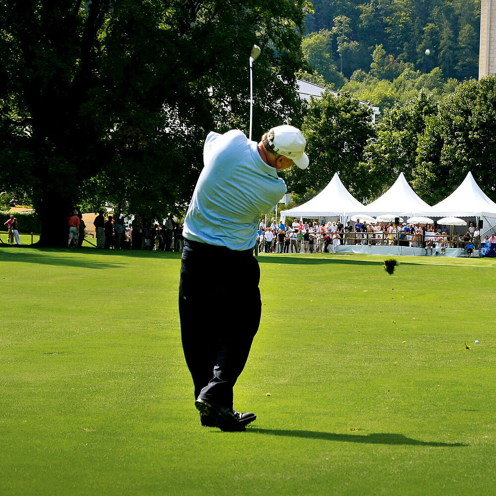Golfer in einem hellblauen Hemd und einer dunklen Hose auf einem begrünten Golfplatz, mitten im Schwung. Weiße Zelte und eine bewaldete Kulisse vervollständigen die sonnige Szene.