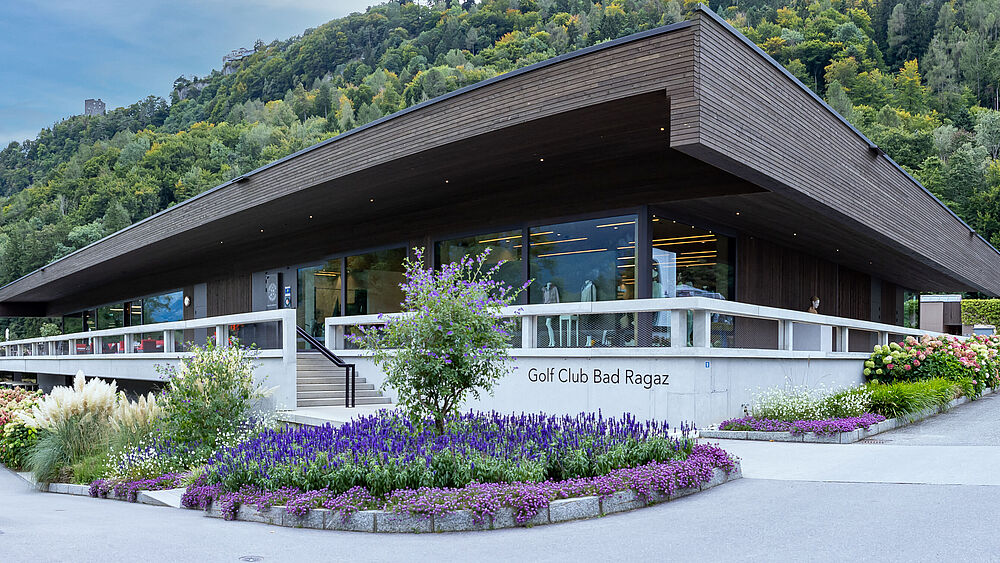 Das Clubhaus des Golf Club Bad Ragaz. Ein modernes Gebäude mit Flachdach, umgeben von Gärten an einem Berghang. Blumen im Vordergrund, Wald im Hintergrund, hell und heiter.