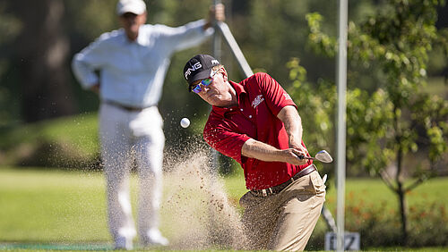 Zwei Golfspieler. Der im Vordergrund trägt ein rotes Shirt und ein dunkles Cap. Er hat soeben den Ball aus dem Bunker geschlagen. Im Hintergrund wartet ein Herr ganz in weiss gekleidet und schaut gespannt zu.
