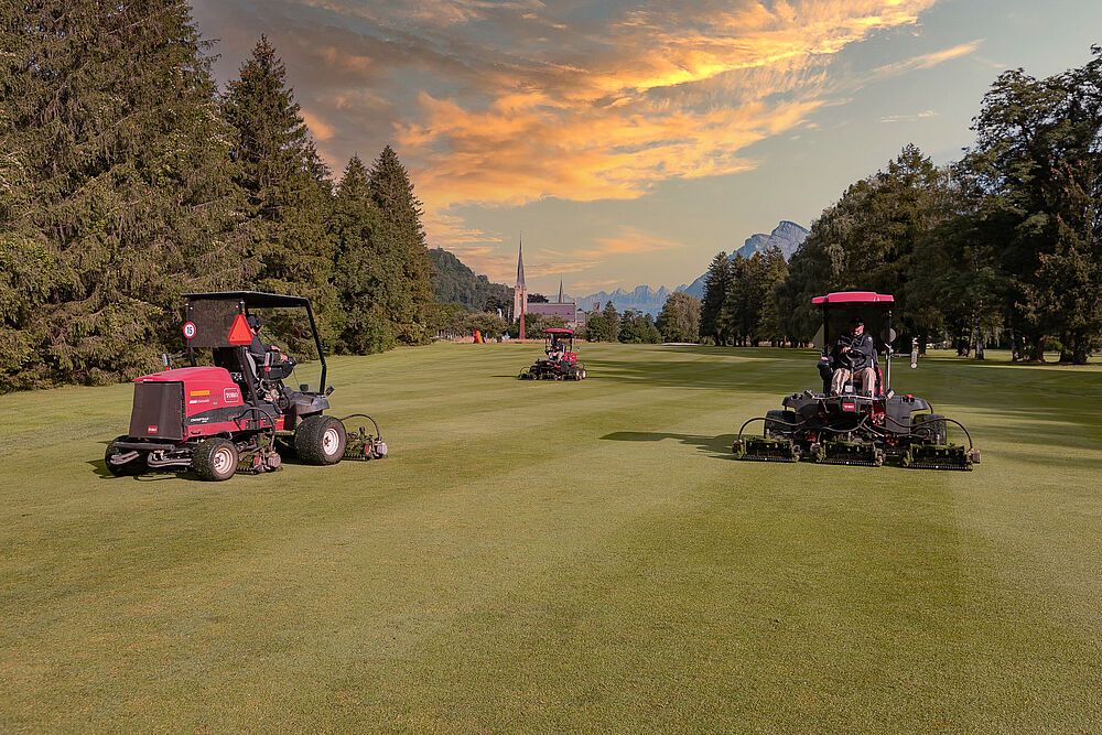 Three red lawnmowers on a huge, lush green field under a dramatic sunset sky. Trees line the field, the fairway, creating a serene and picturesque scene.